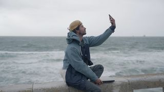 A man sits by the ocean and takes a selfie on a stormy day