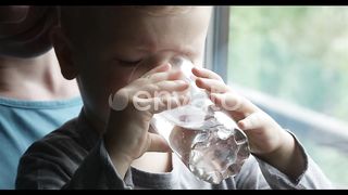 Little Boy Drinking Water From a Glass and Smiling, with Mom Holding Him by the Window