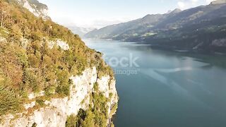 Lakes calm surface meets steep cliff edges covered in dense greenery, framed by distant mountains under a clear blue sky in Switzerland .
