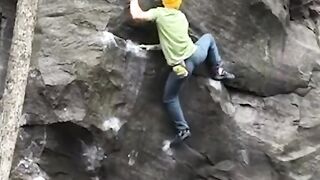 a person wearing a yellow hat, green shirt, and blue jeans is seen bouldering on a large rock face.