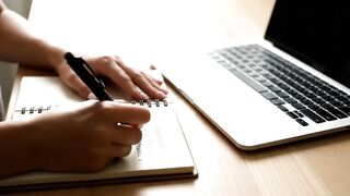 Close-up of Hands Writing in Spiral Notebook Next to Laptop