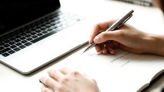 Person Writing in Notebook Next to Laptop Close-up
