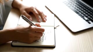 Woman Writing in Notebook Next to Laptop on Desk
