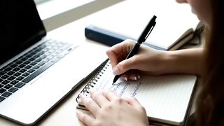 Woman Writing in Notebook Next to Laptop on Desk 2