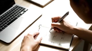 Person Writing in Planner Next to Laptop on Wooden Desk
