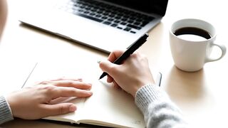 Person Writing in Notebook at Desk with Laptop and Coffee