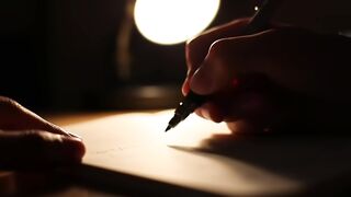 Hand Writing on Paper Under Desk Lamp in Dark Room