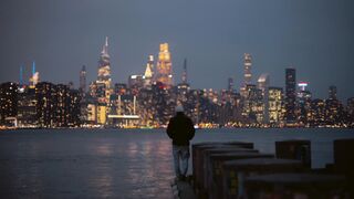 A person stands by the water with NYC skyline illuminated at dusk.