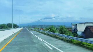 A driver’s perspective on a quiet highway, revealing a view of a volcano, a blue sky with clouds, rural houses, and green trees.